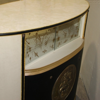 Realizado en polipiel, superficie plástica, madera ebonizada (patas), metal dorado y cristal decorado.